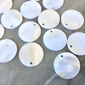 May include: A collection of white, round, mother of pearl shell discs with a small hole in the center. The discs are smooth and have a slight iridescent sheen.