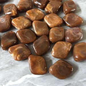 Può includere: Una collezione di perline quadrate marroni con un motivo marmorizzato e a spirale. Le perline hanno una finitura lucida e sono probabilmente realizzate in resina o materiale simile. Ogni perlina ha un foro per l'infilatura.