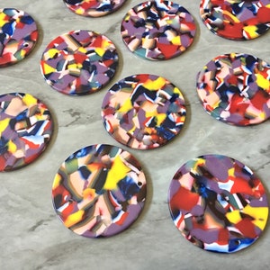 May include: A collection of ten round, colorful, multi-colored, and patterned acrylic resin cabochons. The cabochons are approximately 1.5 inches in diameter and have a hole drilled through the top.