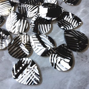 May include: A collection of black, white, and gray teardrop-shaped beads. Each bead features a striped pattern, with a hole at the top for stringing. The beads are scattered on a light gray surface.