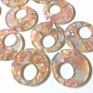 May include: A group of ten round, translucent, pink and gold marbled plastic rings with a hole in the center. The rings are scattered on a white surface.