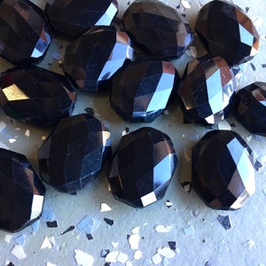 May include: A group of black faceted glass beads, scattered on a textured surface. The beads are oval shaped and have a glossy finish.