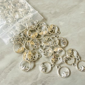 Könnte beinhalten: Eine Tüte mit silbernen Metall-Anhängern mit einem Bergmotiv. Die Anhänger sind rund und haben oben ein Loch zum Anbringen an Schmuck oder andere Bastelarbeiten.