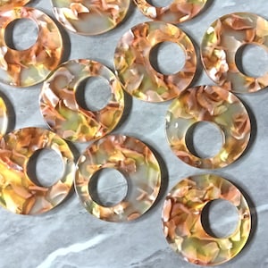 May include: A collection of translucent, circular acrylic beads with a hole in the center. The beads feature a mottled pattern of orange, yellow, and cream colors, resembling a floral or abstract design. These beads are suitable for jewelry making.