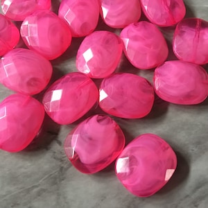 May include: A collection of faceted, hot pink beads with a marbled effect. The beads are oval-shaped with flat sides and are scattered across a gray surface. The beads have a translucent quality, allowing light to pass through.