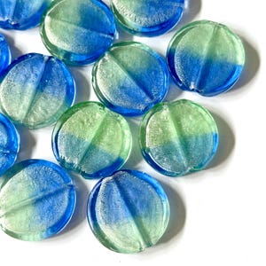May include: A collection of round, glass beads in shades of blue and green. Each bead is divided into two color sections, creating a gradient effect. The beads are arranged on a white surface.