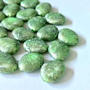 Könnte beinhalten: Eine Sammlung hellgrüner, ovaler Perlen mit einem Craquelé-Effekt. Die Perlen sind auf einer weißen Oberfläche verstreut und zeigen ihre glänzende Oberfläche und komplizierten, netzartigen Muster. Ideal für die Schmuckherstellung.