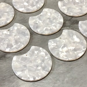 Puede incluir: Una colección de ocho piezas decorativas blancas, semicirculares, iridiscentes, con un agujero en la parte superior. Las piezas están hechas de un material que se asemeja a la madreperla.