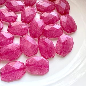 May include: A collection of faceted, pink gemstone beads. Each bead has a geometric shape with multiple flat surfaces, creating a sparkling effect. The beads are arranged on a white surface, showcasing their vibrant color and texture.