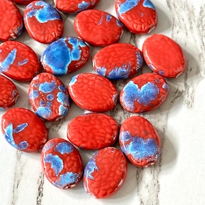 May include: Oval-shaped ceramic beads in a vibrant red hue, accented with patches of blue. The beads have a textured surface and are suitable for jewelry making or craft projects. The beads are approximately 1 inch in length.