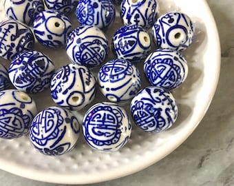 May include: A selection of round, white ceramic beads featuring detailed blue patterns. The beads are presented on a white plate, highlighting the contrast between the blue and white. Each bead has a hole for threading, showcasing the intricate designs.