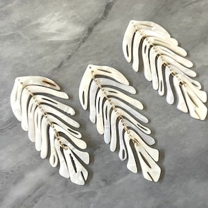 Puede incluir: Tres pendientes blancos en forma de hoja con detalles de cadena dorada. Los pendientes están hechos de un material nacarado y tienen un diseño delicado y orgánico.