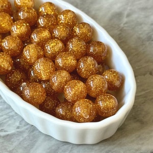 May include: A white bowl filled with gold glitter beads. The beads are round and have a shiny finish.