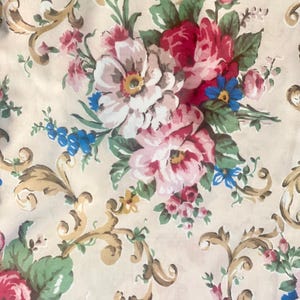 Op de afbeelding: Een beige stof met een bloemenpatroon. Het patroon bevat roze, rode en blauwe bloemen met gouden accenten. De bloemen zijn gerangschikt in een herhalend patroon.