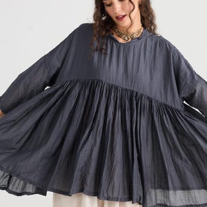 May include: A dark gray, flowing top with a gathered waist and wide sleeves. The top has a high neckline and a loose, comfortable fit. The model is wearing a gold necklace and light-colored pants.
