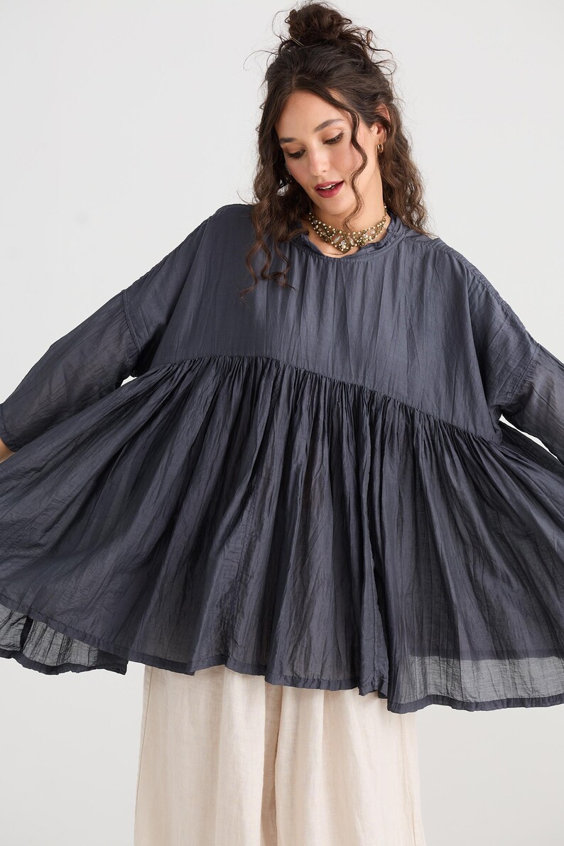 May include: A dark gray, flowing top with a gathered waist and wide sleeves. The top has a high neckline and a loose, comfortable fit. The model is wearing a gold necklace and light-colored pants.