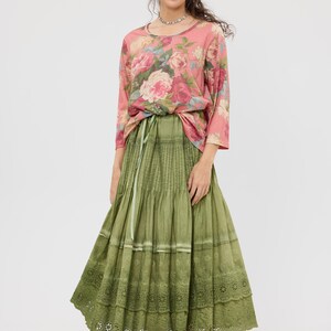 Puede incluir: Una mujer con una blusa rosa con estampado floral y mangas tres cuartos, y una falda larga verde oliva. La falda tiene un dise&ntilde;o escalonado con un dobladillo de encaje. El conjunto se ata a la cintura con una cinta verde. Lleva un collar plateado y bailarinas rosas.
