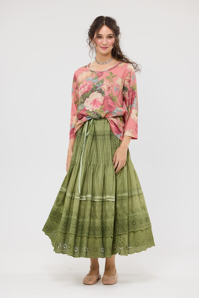 Puede incluir: Una mujer con una blusa rosa con estampado floral y mangas tres cuartos, y una falda larga verde oliva. La falda tiene un dise&ntilde;o escalonado con un dobladillo de encaje. El conjunto se ata a la cintura con una cinta verde. Lleva un collar plateado y bailarinas rosas.