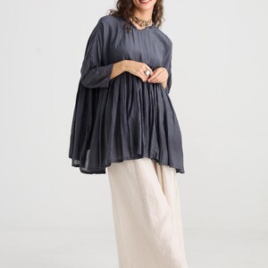 May include: A dark gray tunic top with a round neckline and three-quarter sleeves. The top is paired with cream-colored wide-leg pants. The outfit is completed with black flats. The clothing is made of a soft, flowing fabric.
