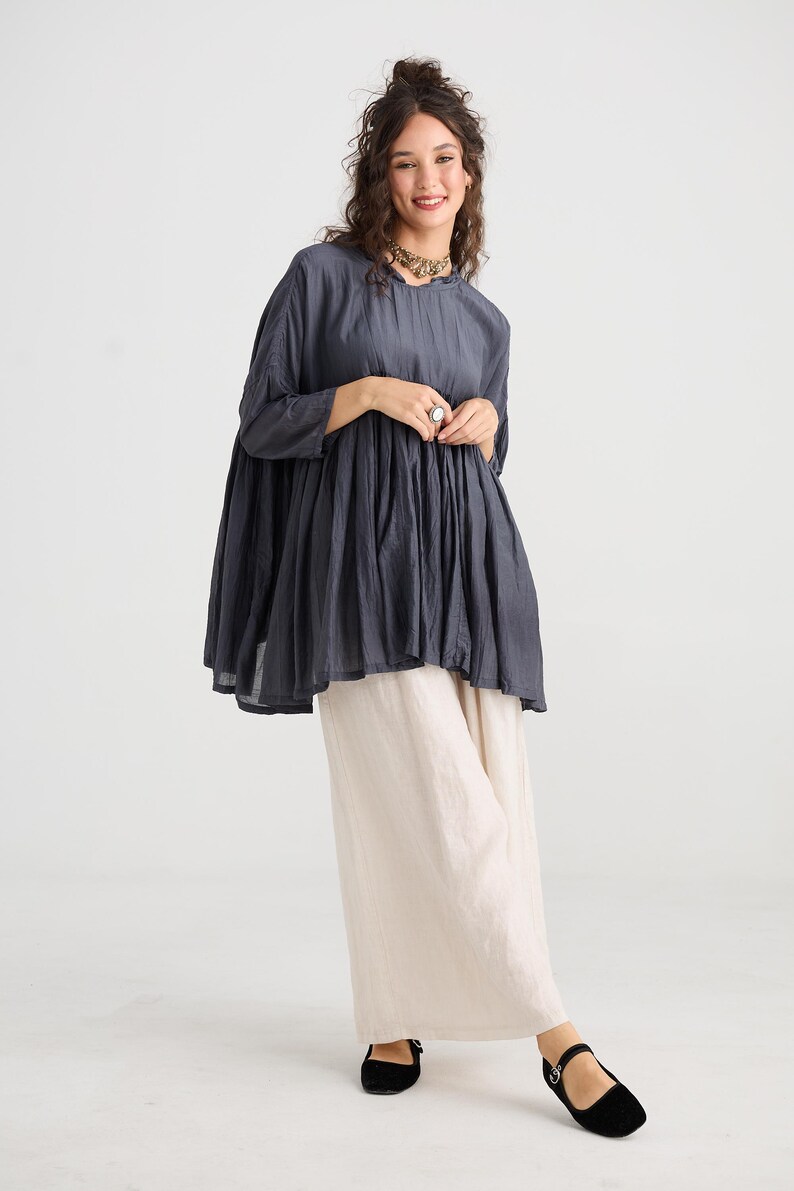 May include: A dark gray tunic top with a round neckline and three-quarter sleeves. The top is paired with cream-colored wide-leg pants. The outfit is completed with black flats. The clothing is made of a soft, flowing fabric.