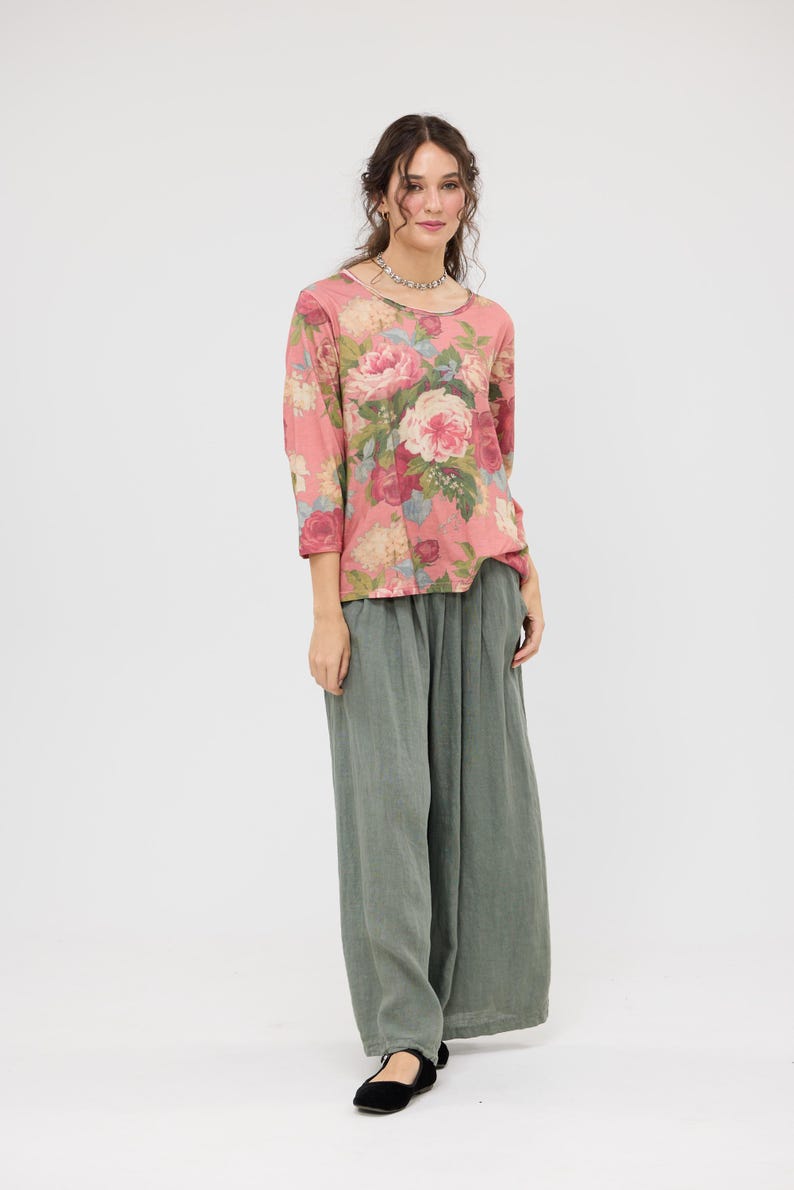 Puede incluir: Una blusa rosa con estampado floral, mangas tres cuartos y cuello redondo. Combinada con pantalones de pierna ancha verde oliva y bailarinas negras. La blusa presenta un gran estampado de rosas en tonos rosa, rojo y crema.