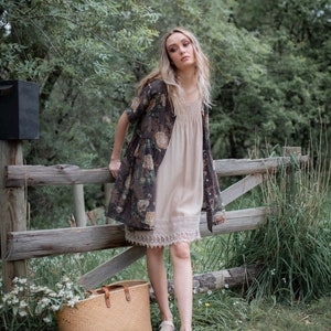 Puede incluir: Una mujer con un vestido de estampado floral con ribete de encaje y un c&aacute;rdigan marr&oacute;n se encuentra frente a una valla de madera. Lleva un bolso de mimbre y mira a la c&aacute;mara.