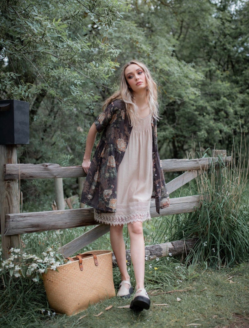 Puede incluir: Una mujer con un vestido de estampado floral con ribete de encaje y un c&aacute;rdigan marr&oacute;n se encuentra frente a una valla de madera. Lleva un bolso de mimbre y mira a la c&aacute;mara.