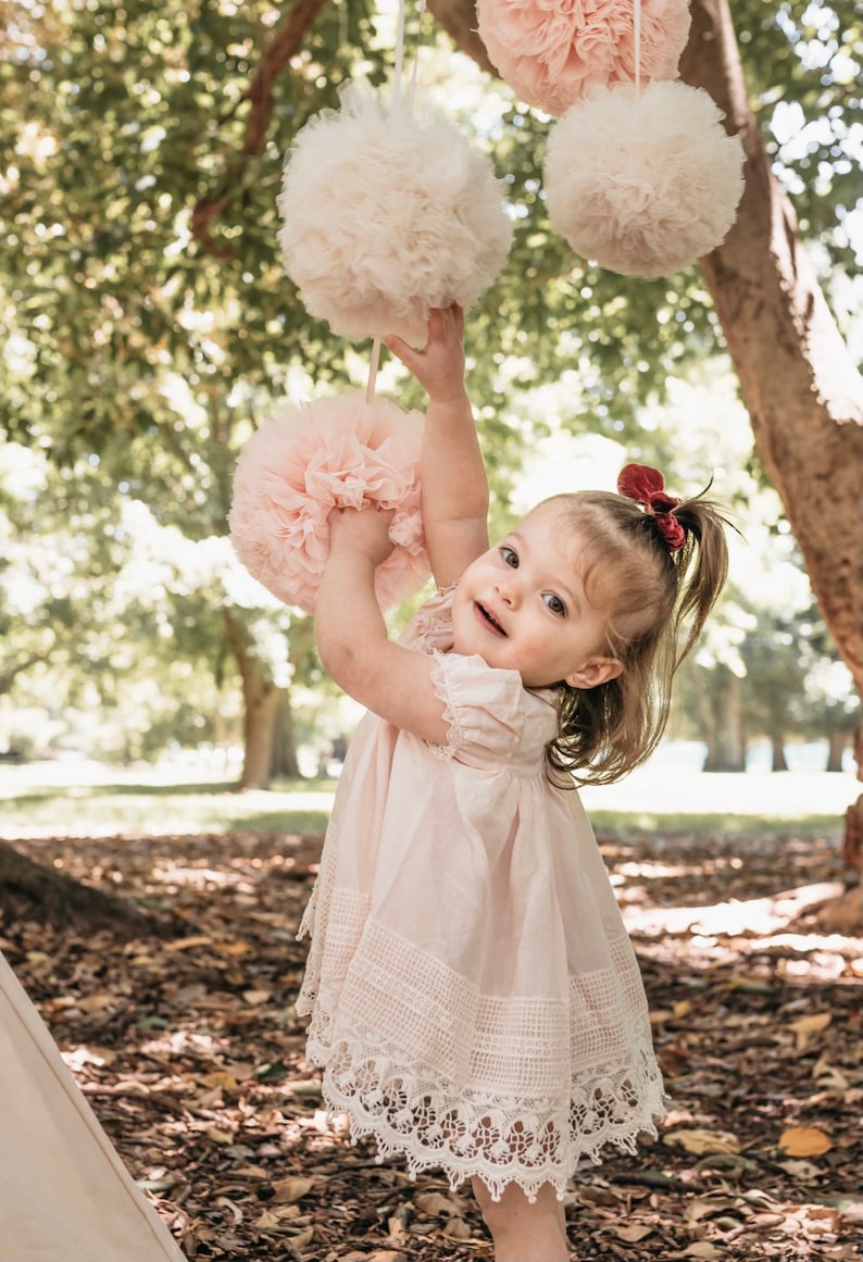 May include: A young girl in a pink dress with lace trim reaches up to touch a pink tulle pom-pom hanging from a tree branch. There are other pink tulle pom-poms hanging in the background.
