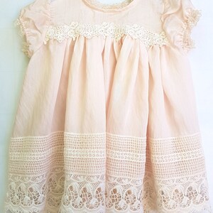 May include: A pale pink baby dress with delicate white lace trim around the neckline, sleeves, and hem. The dress has a gathered skirt and a simple, classic design.