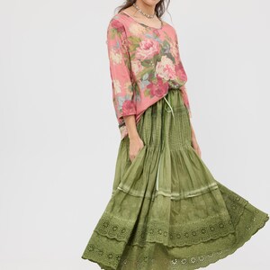Puede incluir: Una mujer con una blusa rosa con estampado floral y una falda verde oliva escalonada. La falda tiene un ribete de encaje en el dobladillo. El atuendo es de estilo bohemio.