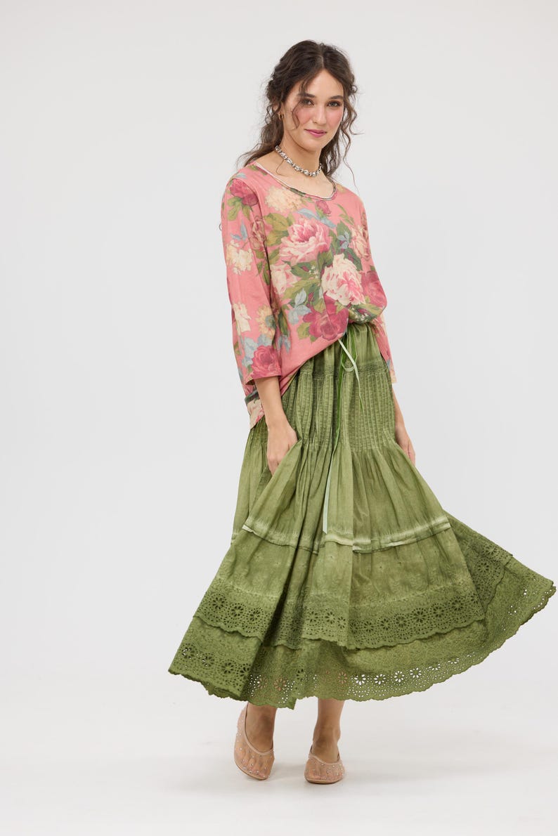 Puede incluir: Una mujer con una blusa rosa con estampado floral y una falda verde oliva escalonada. La falda tiene un ribete de encaje en el dobladillo. El atuendo es de estilo bohemio.