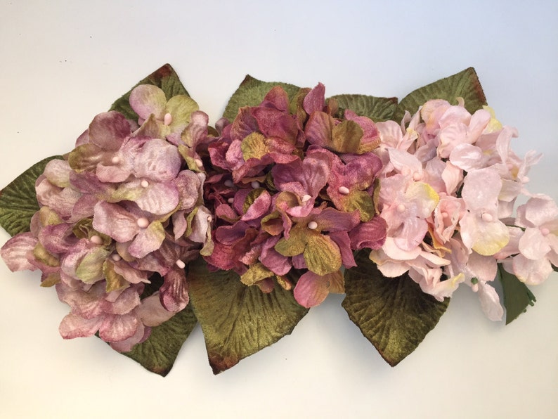 May include: Three artificial hydrangea flower heads in shades of pink, purple, and green. The flowers are arranged in a row and have velvet-like leaves.