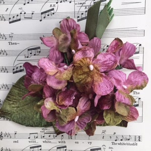 May include: A cluster of pink and green velvet flowers, resembling hydrangeas, arranged on a sheet of music. The music sheet features the words "wandered in de light" and "The white blush'd ten."