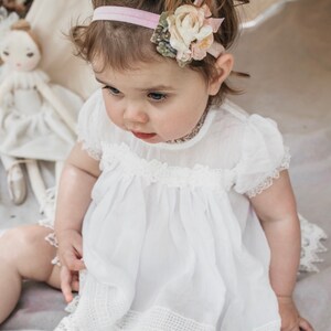 May include: A baby girl wearing a white dress with lace trim and a pink headband with a flower accent.
