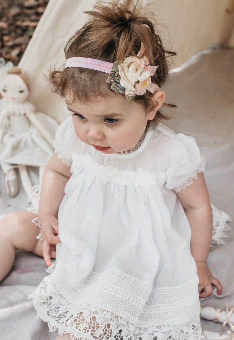 May include: A baby girl wearing a white dress with lace trim and a pink headband with a flower accent.