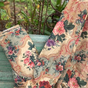 Puede incluir: Una pieza de tela con fondo color crema y un patr&oacute;n repetitivo de ramos de flores rosas, rojas y moradas. El dise&ntilde;o incluye bordes ornamentados de color beige y finas rayas verticales de color verde oscuro entre los motivos.