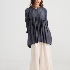 May include: A woman wearing a dark gray tunic top with long sleeves and a gathered bodice, paired with cream-colored wide-leg pants. She is also wearing a gold necklace and black flats. The outfit is simple and elegant.
