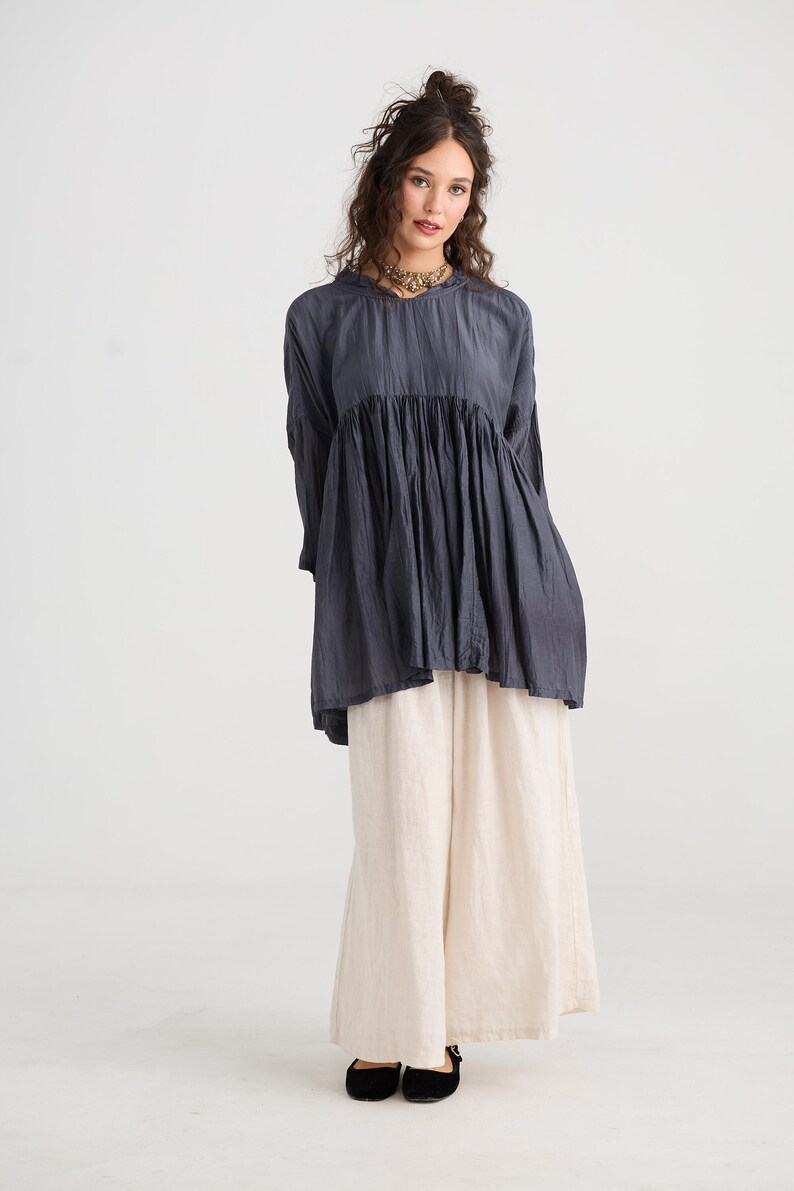 May include: A woman wearing a dark gray tunic top with long sleeves and a gathered bodice, paired with cream-colored wide-leg pants. She is also wearing a gold necklace and black flats. The outfit is simple and elegant.