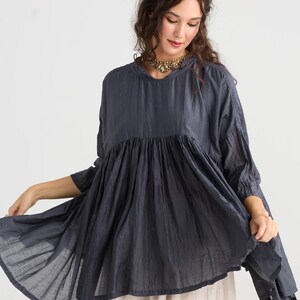 May include: A dark blue tunic top with a gathered bodice and flared sleeves. The top is paired with a light beige skirt. The model is wearing a gold-toned necklace.