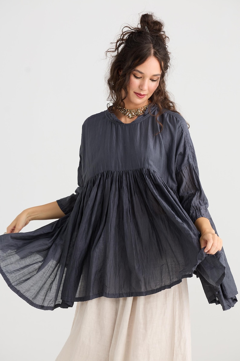 May include: A dark blue tunic top with a gathered bodice and flared sleeves. The top is paired with a light beige skirt. The model is wearing a gold-toned necklace.