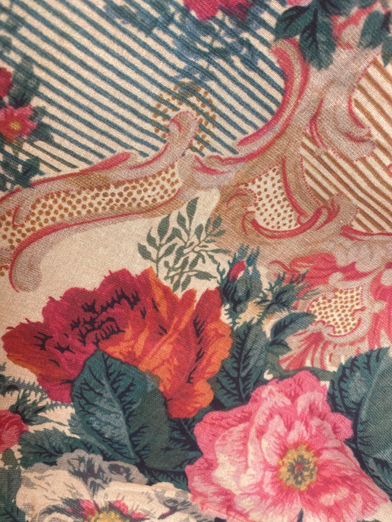 Puede incluir: Una tela con un estampado floral de grandes flores rojas, rosas y blancas, hojas verdes y elementos decorativos en color tostado. El fondo consiste en finas rayas diagonales alternas en azul y blanco, creando un efecto visual.