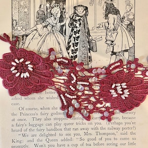 May include: A close-up of a red and white lace trim with white beads and sequins. The lace is on top of a page of a book with text that reads "Of course, when she", "the Princess's fairy godmother", "at once. They also stopped offering", "the nursery", "case, because", "a fairy's luggage can play queer tricks on you."