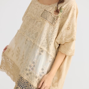 May include: Beige, oversized tunic top with a round neckline and short sleeves. The top features intricate lace and eyelet details, with a patchwork design. A small floral embellishment and a patch of text are visible on the front. The bottom hem has a crochet design.