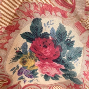 Puede incluir: Una tela que presenta un ramo floral central con flores rojas, rosas, amarillas y moradas rodeadas de hojas verde oscuro. El fondo consiste en un panel de color crema enmarcado por adornos rosas y patrones de rayas de color tostado.