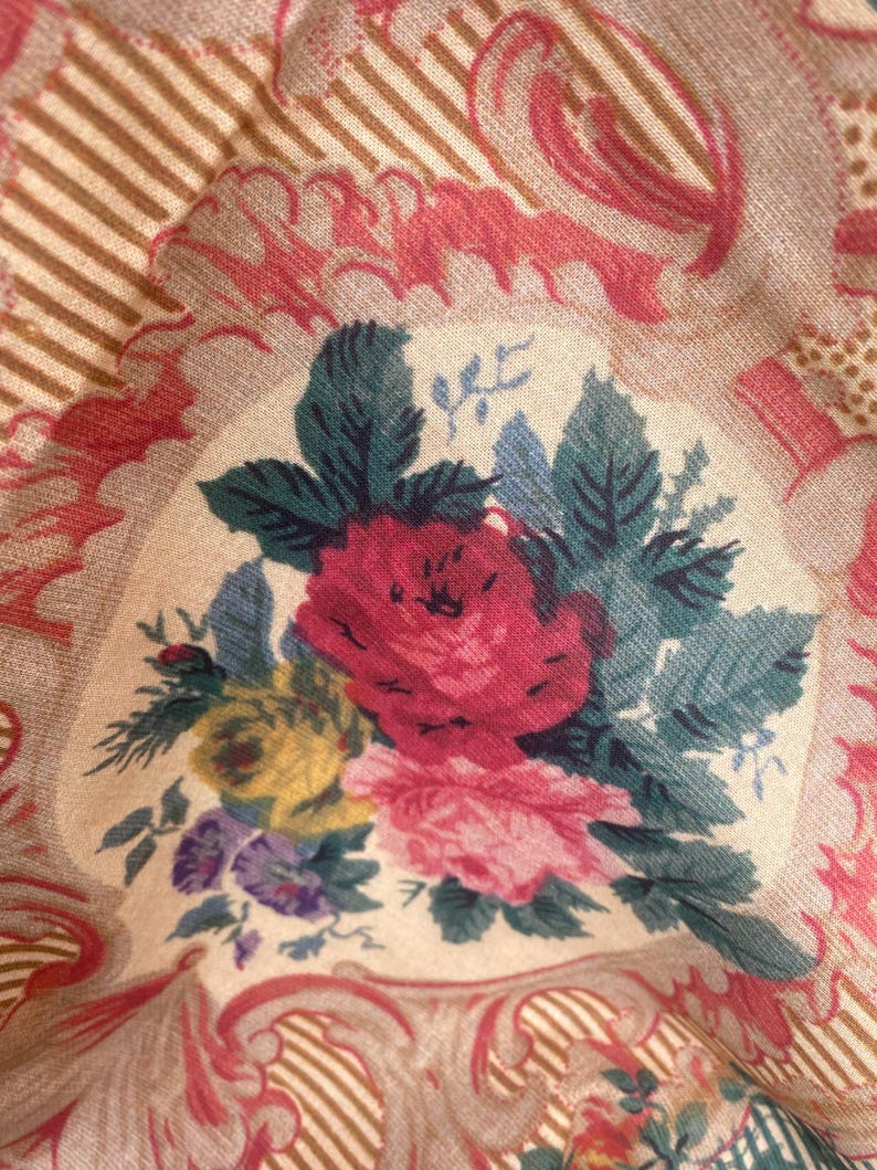 Puede incluir: Una tela que presenta un ramo floral central con flores rojas, rosas, amarillas y moradas rodeadas de hojas verde oscuro. El fondo consiste en un panel de color crema enmarcado por adornos rosas y patrones de rayas de color tostado.