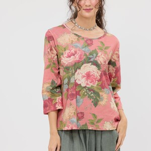 Puede incluir: Una blusa rosa con estampado floral y mangas tres cuartos. La blusa presenta un cuello redondo y un vibrante estampado de rosas rosas y blancas con follaje verde. La modelo lleva un collar plateado y pantalones verdes.