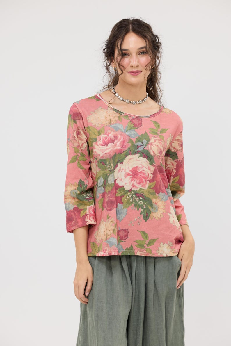 Puede incluir: Una blusa rosa con estampado floral y mangas tres cuartos. La blusa presenta un cuello redondo y un vibrante estampado de rosas rosas y blancas con follaje verde. La modelo lleva un collar plateado y pantalones verdes.