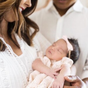 May include: A couple holding a sleeping baby wearing a white crocheted dress with a pink bow. The baby is being held by the father, while the mother looks down at the baby.
