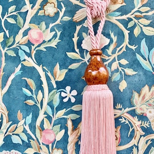 May include: A pink tassel tieback with a wooden finial. The tieback is hanging against a blue and green floral patterned fabric.