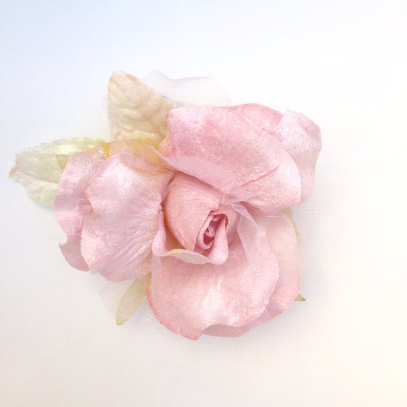 May include: A delicate pink fabric rose with a soft, velvety texture. The rose is made of multiple layers of petals and has a slightly ruffled appearance.